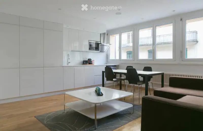 Spacious open-plan living room and kitchen with modern, minimalist design. The kitchen features white cabinets and appliances, including a dishwasher. The living area has a large white coffee table and dining table with black chairs. The room is well-lit