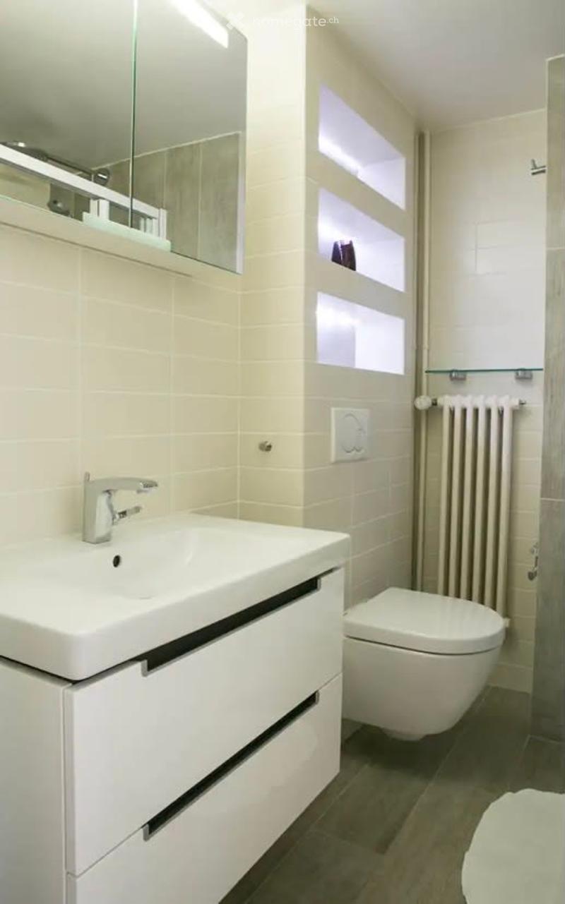 Modern bathroom with white vanity, sink, toilet, and tiled walls. There is a radiator and a window providing natural light.