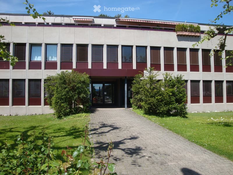 Modern two-story building with a flat roof, large windows, and a central entrance. The building has a symmetrical design with a central pathway leading to the entrance, surrounded by well-maintained grass and shrubs.