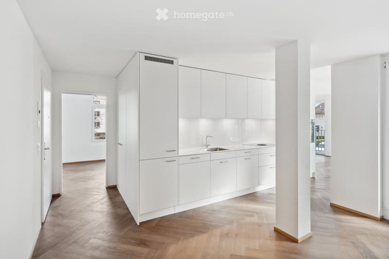 Modern, minimalist kitchen with white cabinets, a central island, and a sink. The space features light wooden flooring and ample natural light from a window.