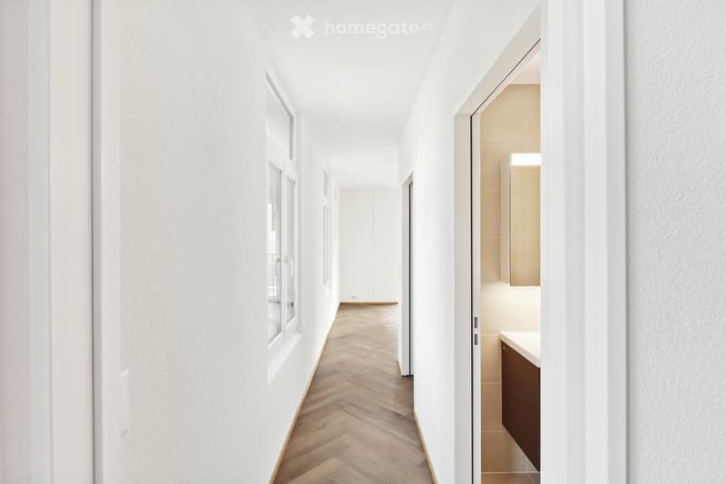 A modern, minimalist hallway with white walls, herringbone wooden flooring, and several windows on one side. A small bathroom is visible at the end of the hallway, featuring a white countertop and dark cabinetry.