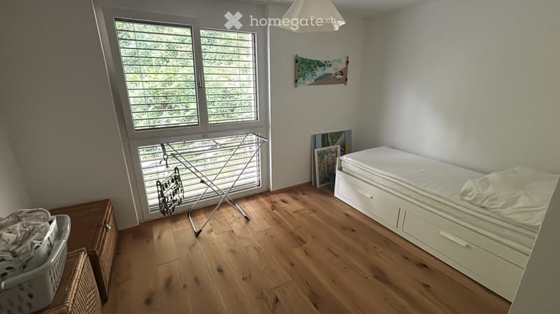 A small bedroom with a wooden floor, a bed with white bedding, a laundry drying rack, a wicker basket, and a laundry basket near the window.