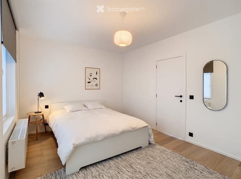 Minimalist bedroom with white walls, wooden flooring, a white bed, nightstand, lamp, and a mirror on the wall.