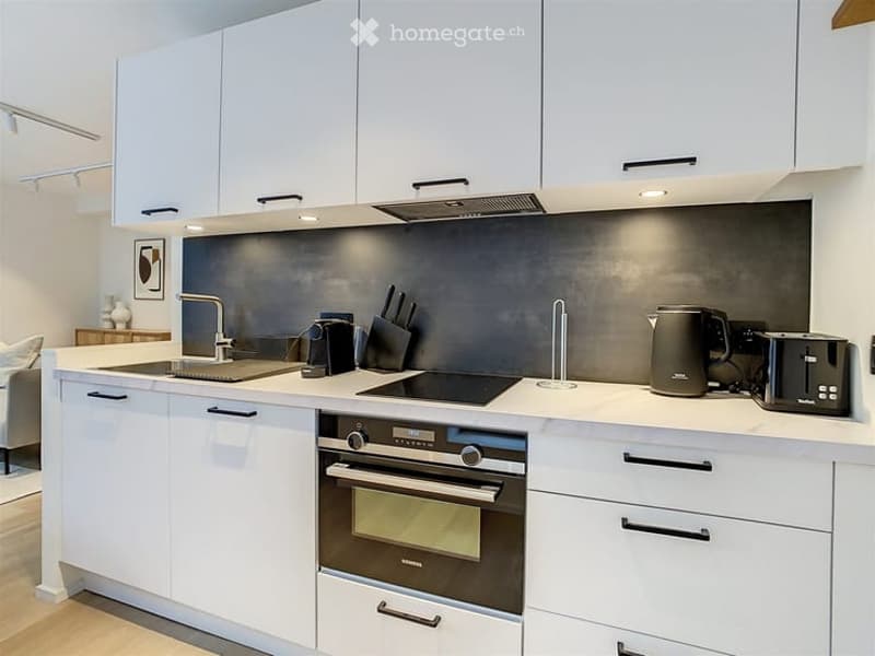 Modern kitchen with white cabinets, stainless steel appliances, and a sleek black backsplash. Features include an induction cooktop, built-in oven, and various small appliances on the counter.