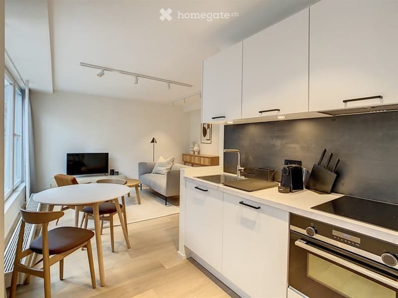 Modern, open-plan living and dining area with a kitchen. Features a grey sofa, coffee table, TV, dining table with chairs, and a sleek kitchen with white cabinets, stainless steel appliances, and a countertop with a blender and knives.