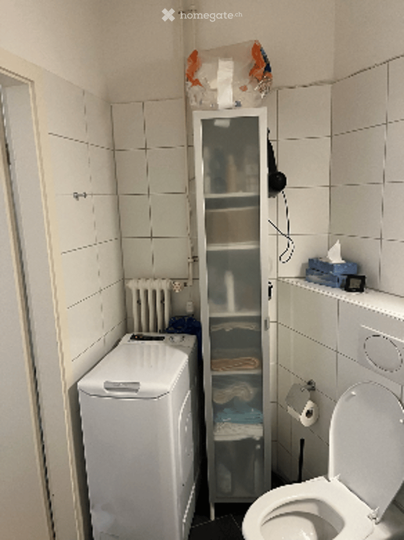 Small bathroom with white tiled walls and floor, featuring a toilet, washing machine, shower cabinet, radiator, and a towel hanging on the wall.