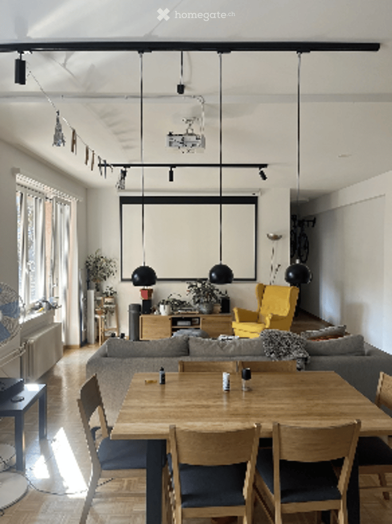 Bright living room with large windows, grey sofa, coffee table, dining table with chairs, pendant lights, and a plant in the corner.