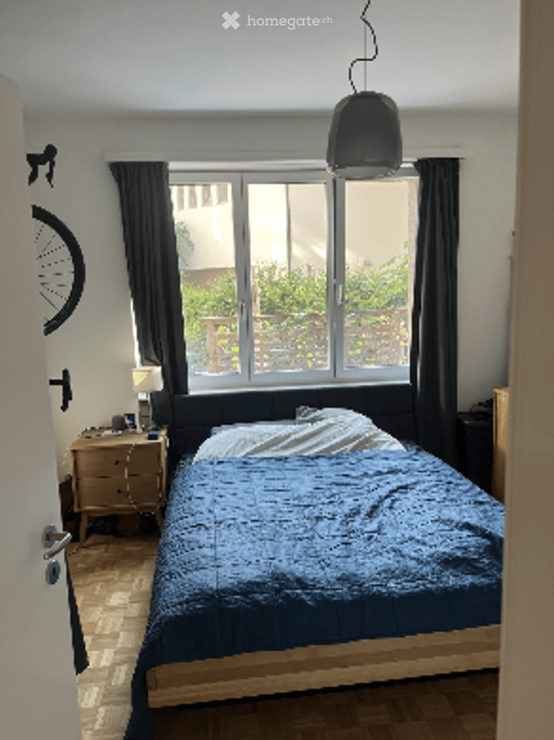 Cozy bedroom with a large window offering a view, a neatly made bed with blue bedding, a nightstand with a lamp, and a mirror on the wall.
