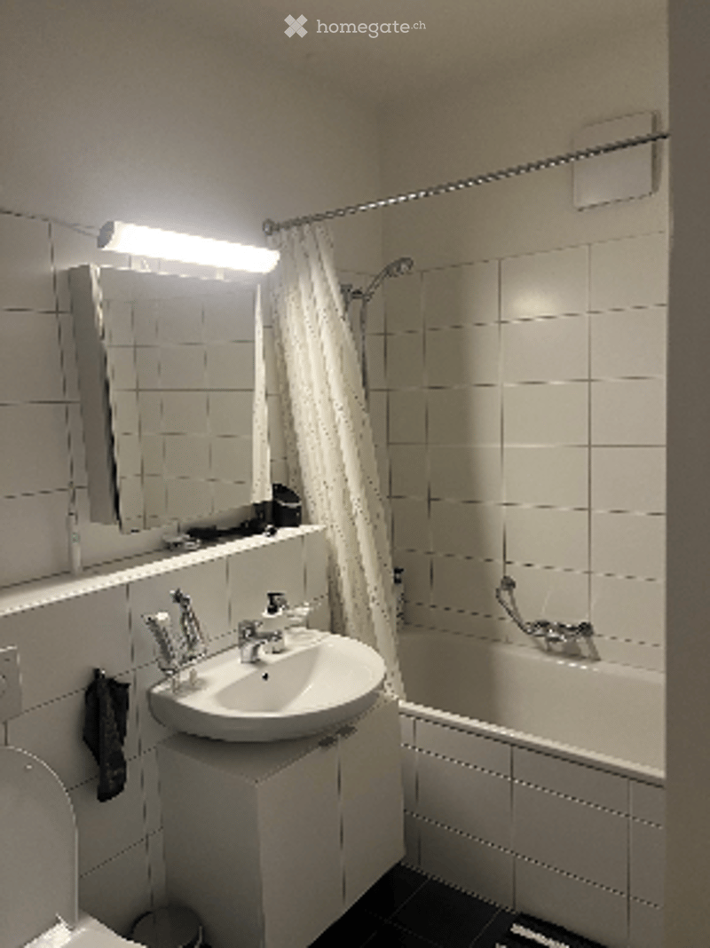 Modern bathroom with white tiles, a sink with a mirror above it, a bathtub with a shower curtain, and a light fixture above the mirror.