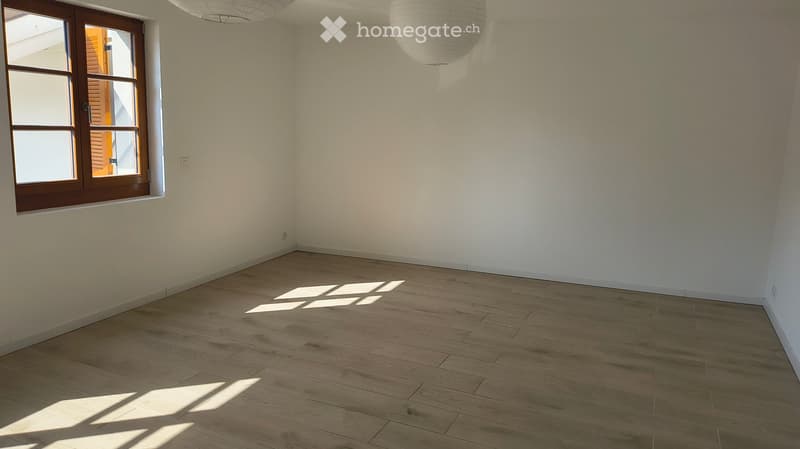 Empty room with light wooden flooring, white walls, and a large window with wooden frame allowing natural light to enter.