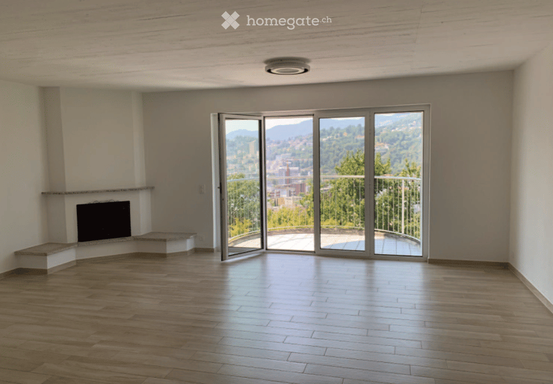 Empty room with wooden flooring, fireplace, large glass doors leading to a balcony with scenic view.