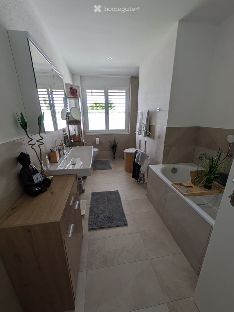 Modern bathroom with a double sink, bathtub, large mirror, and wooden stool. Features include tiled floors, natural light from windows, and decorative plants.