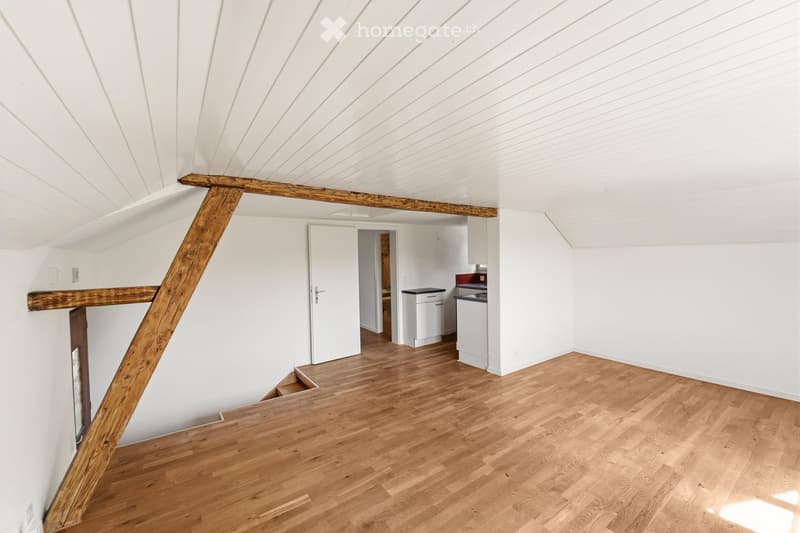 An empty attic space with wooden floors, white walls, and exposed wooden beams. A small kitchen area is visible in the background, but no furniture is present.