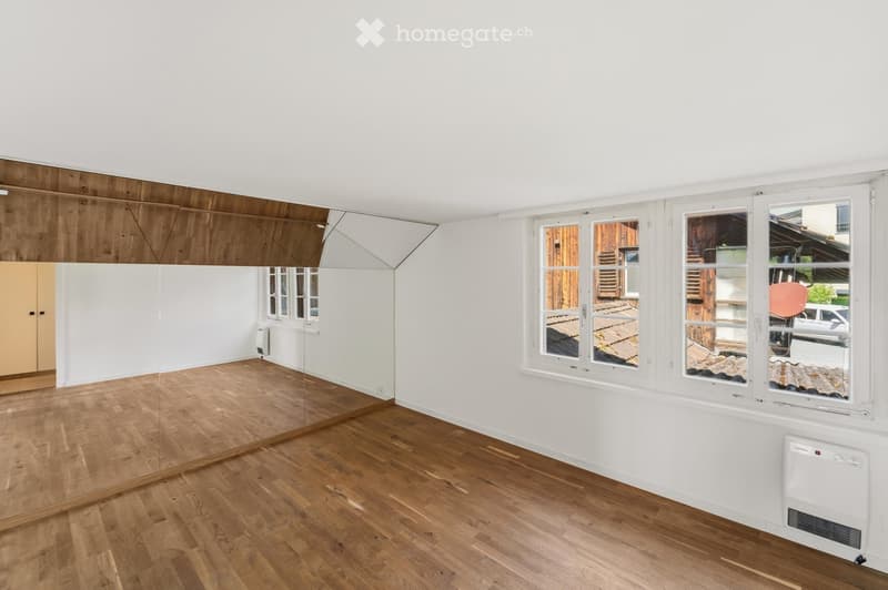 Completely empty room with wooden flooring, white walls, large windows, and a mirrored wall section.