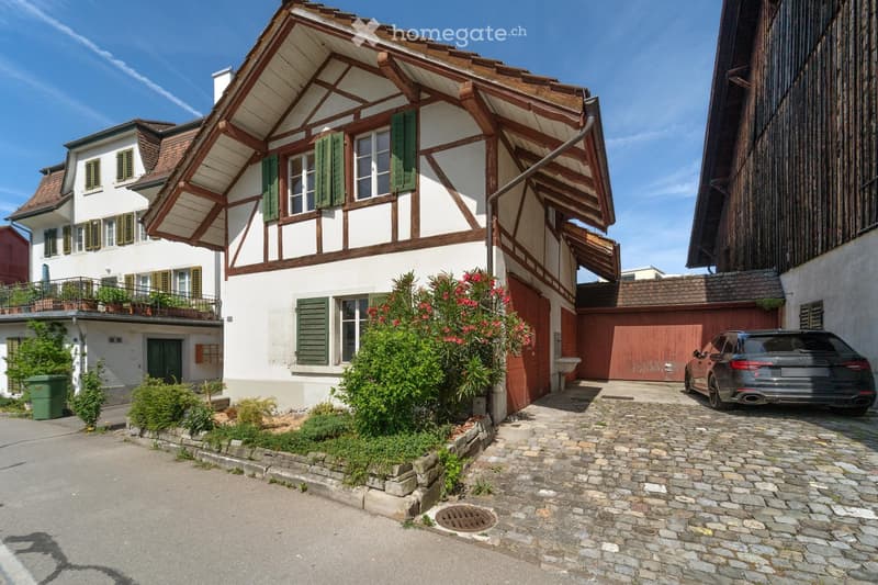 Traditional two-story house with white walls, wooden beams, green shutters, and a red garage door. The house features a balcony with potted plants and is surrounded by small shrubs and flowers. A black car is parked next to the garage.