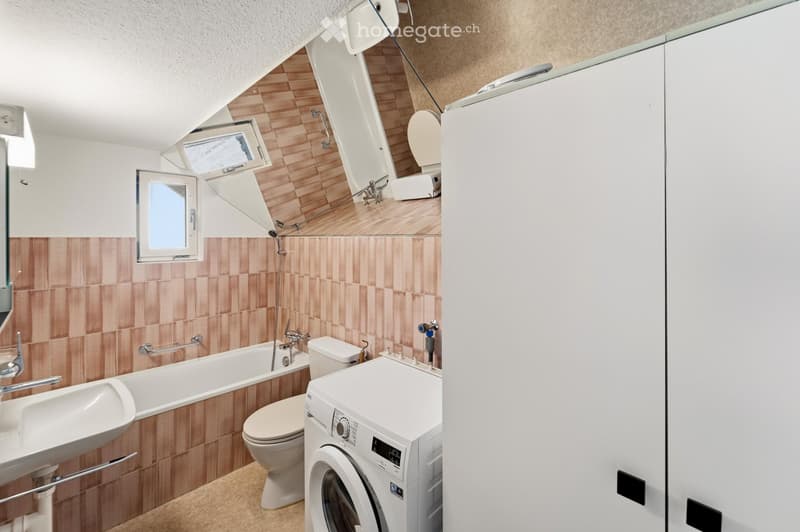 Modern bathroom with a bathtub, toilet, sink, washing machine, and a large mirror. The walls are tiled in a warm, earthy tone, and there is a window providing natural light.