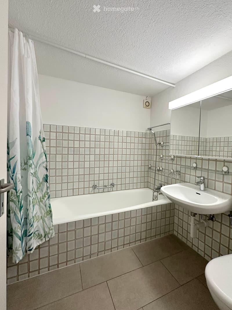 Bathroom with a bathtub, sink, mirror, and shower curtain. The walls are tiled with small square tiles, and the floor is tiled with larger rectangular tiles.