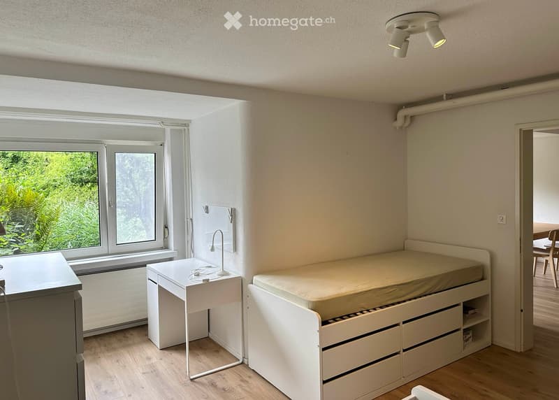 Minimalist bedroom with a single bed, desk, and chair. Large window offers a view of greenery. The room features light-colored walls and wooden flooring.