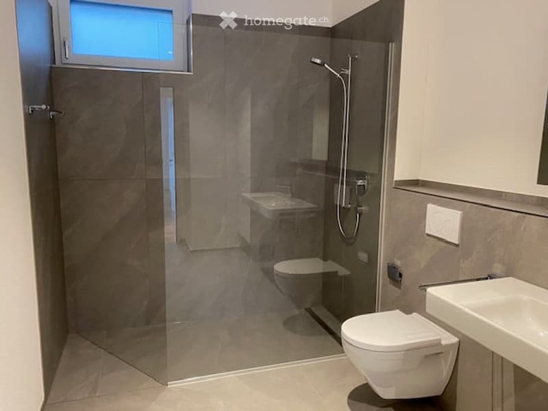 Modern bathroom with a glass-enclosed shower, toilet, and sink. The walls are tiled in a neutral gray color, and the floor is tiled in a light beige color. There is a small window above the shower for natural light.