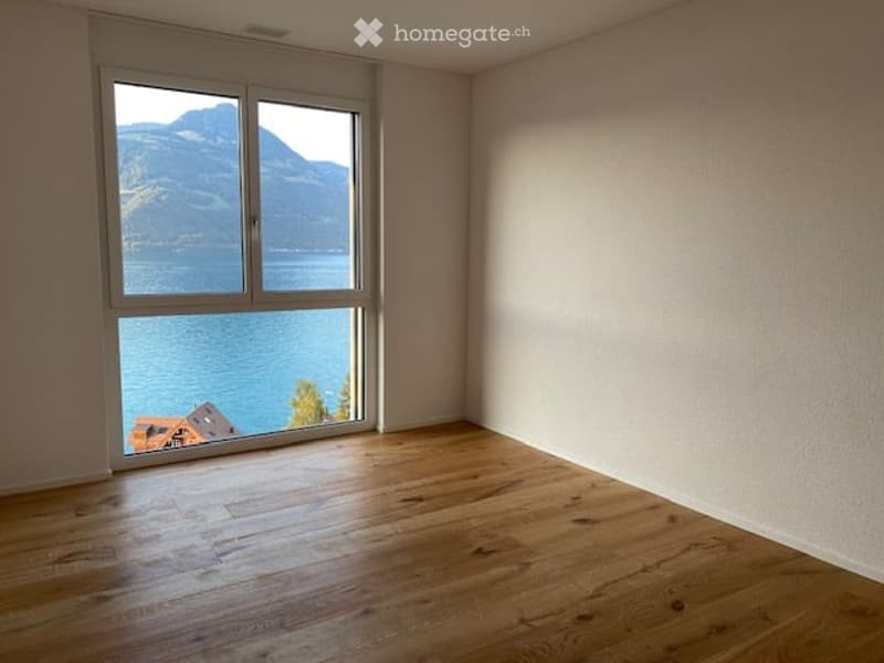 Empty room with wooden flooring and large window offering a scenic view of a lake and mountains.