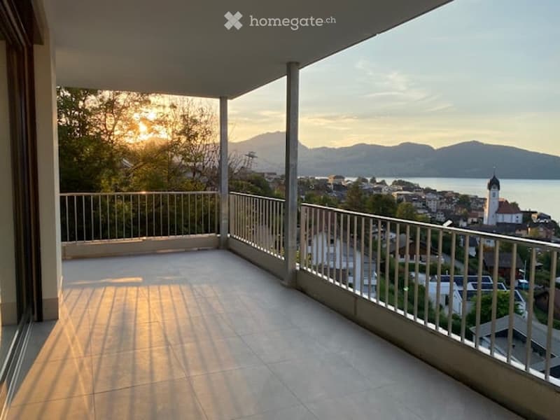 Balcony with a scenic view of a town, lake, and mountains during sunset.