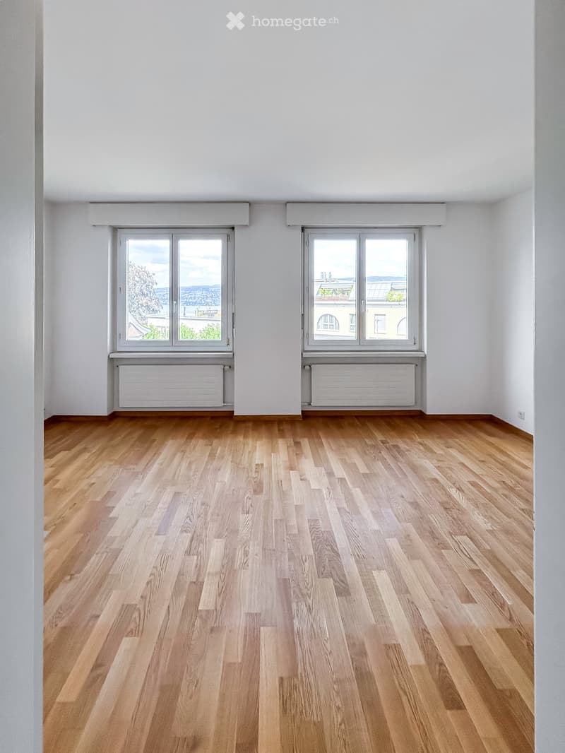 Empty room with wooden flooring, white walls, and two large windows offering a view of the cityscape.