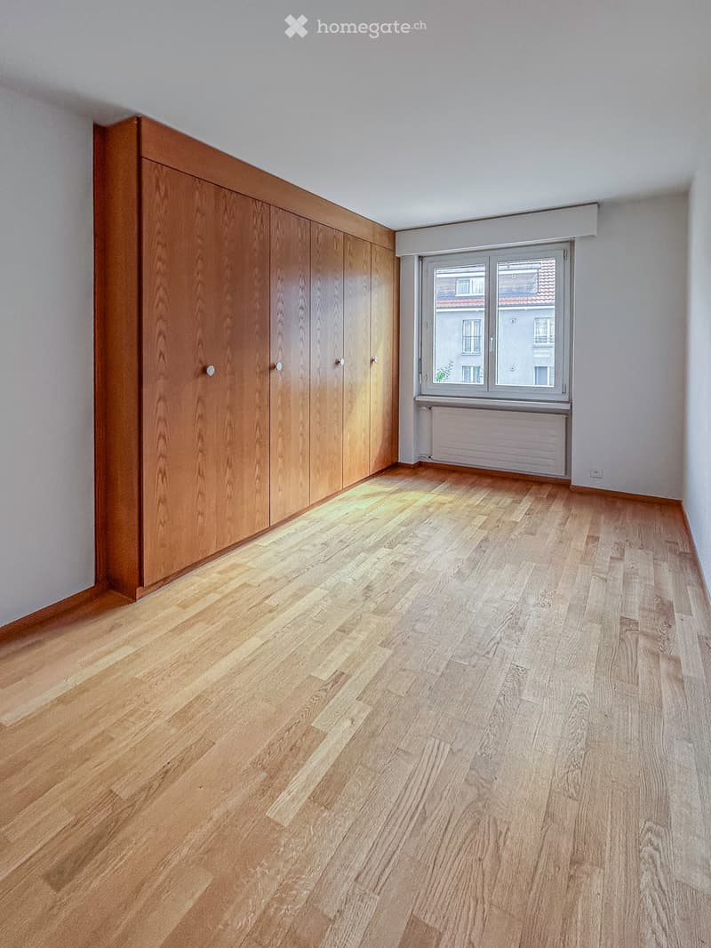 Empty room with wooden flooring, large built-in wardrobe, and a window with a radiator below it.