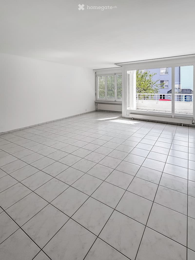 Empty room with grey tiled floor and white walls, featuring large windows and a sliding glass door leading to a balcony.