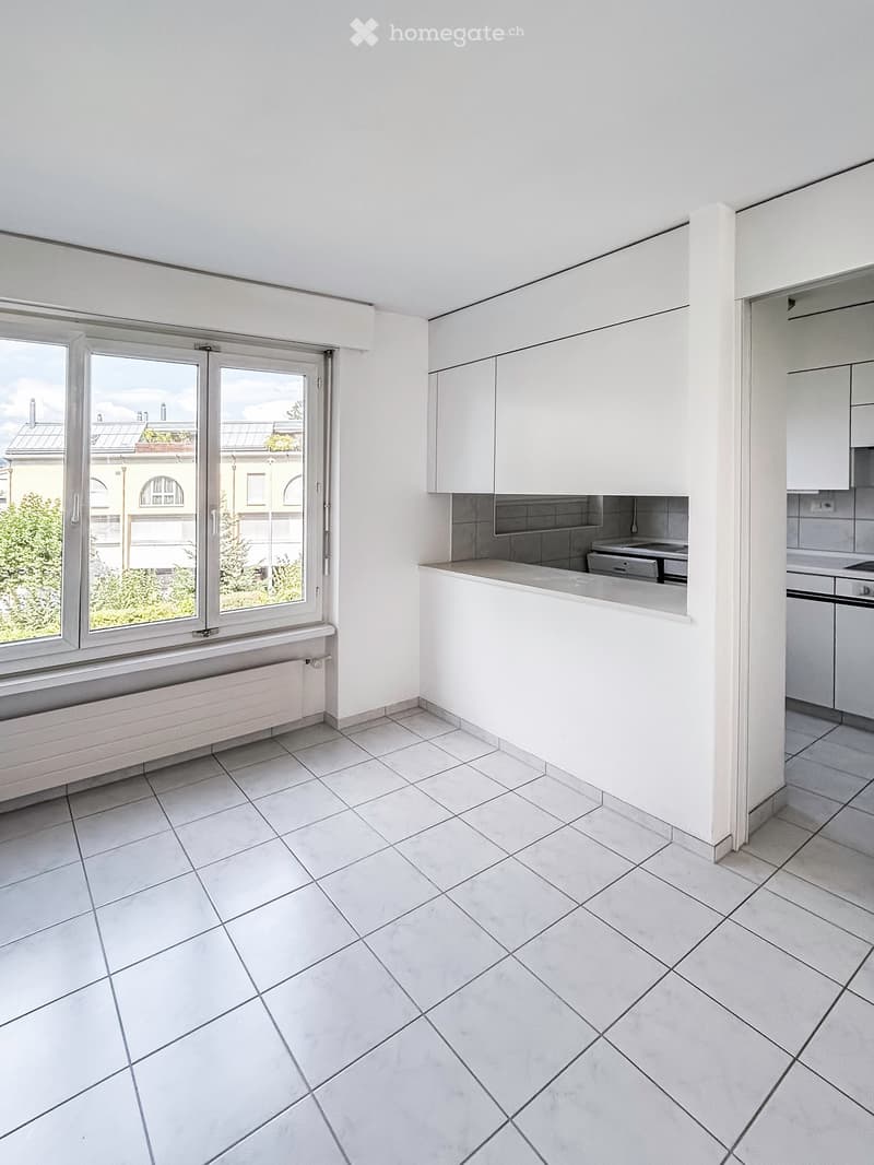 Empty kitchen with white tiled floor, white cabinets, and a window overlooking a cityscape.