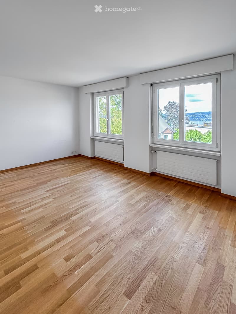 Empty room with wooden flooring, white walls, and large windows offering a view of greenery and buildings.