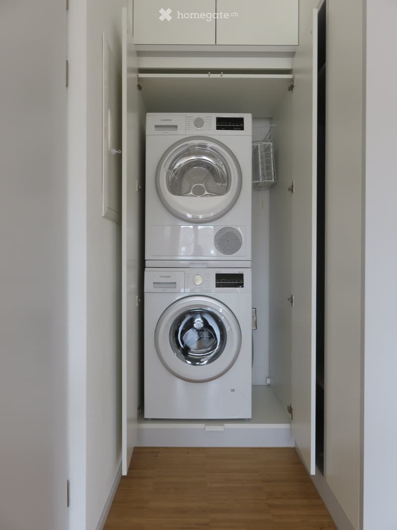 Compact laundry area with stacked Siemens washing machine and dryer, white cabinets, wooden floor.