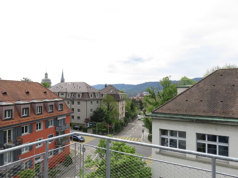 Balcony overlooking a street lined with multi-story residential buildings, featuring red-tiled roofs and various architectural styles, with visible greenery and distant hills.