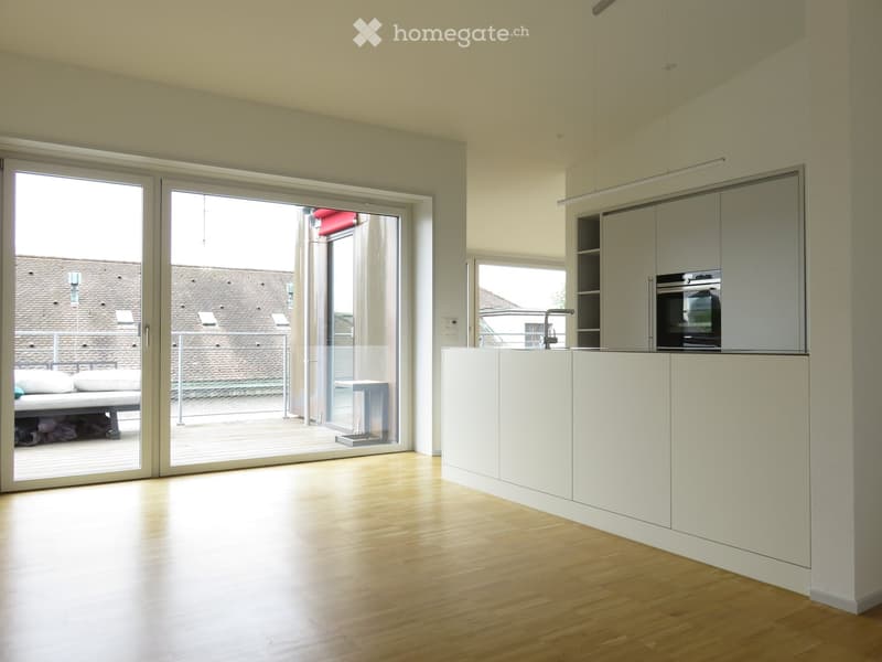 Modern, minimalist room with light wooden flooring, white cabinetry, and a sleek oven. Large sliding glass doors open to a balcony with a view of a tiled roof.