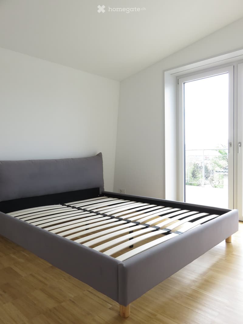 Minimalist bedroom with a grey upholstered bed frame and slatted base, wooden flooring, and a large window with a sliding door leading to a balcony.