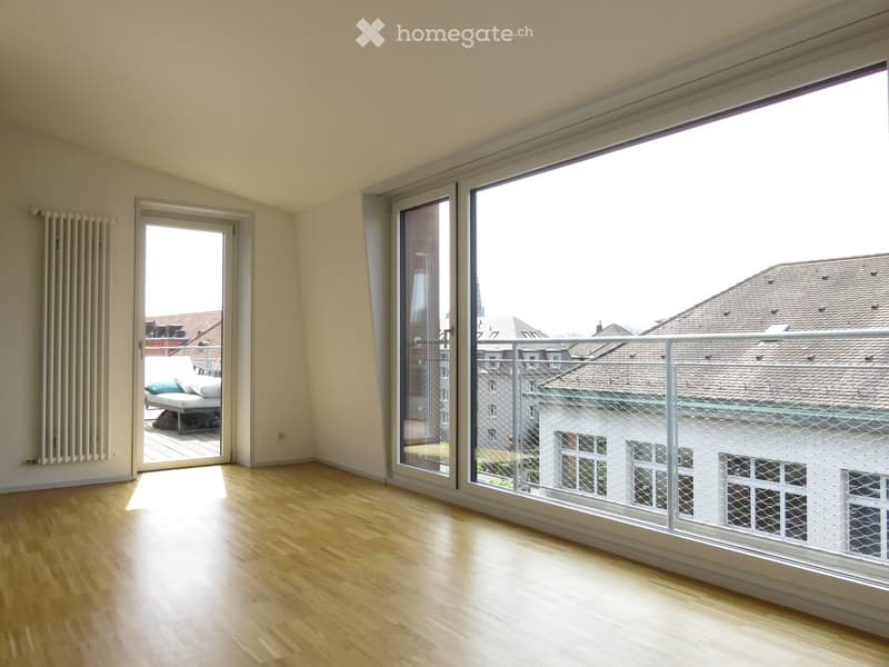 Empty room with wooden flooring, large windows, and a balcony with a metal railing overlooking rooftops.