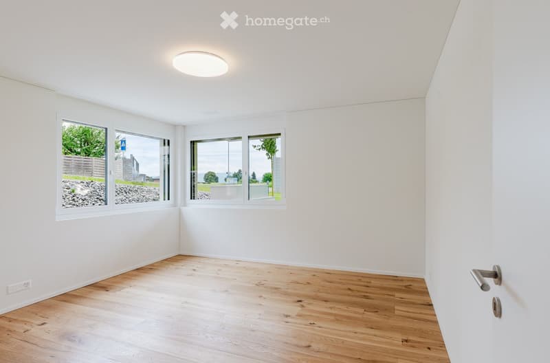 Empty room with white walls, wooden floor, large windows, and a round ceiling light fixture.