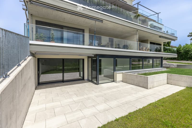 Modern multi-story building with large glass windows and balconies, featuring a raised ground floor and a landscaped garden area.