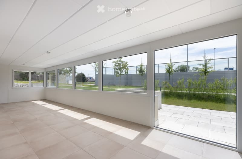 Bright, empty room with large windows and a tiled floor, allowing natural light to fill the space. The room has a modern, minimalist design with white walls and a high ceiling.