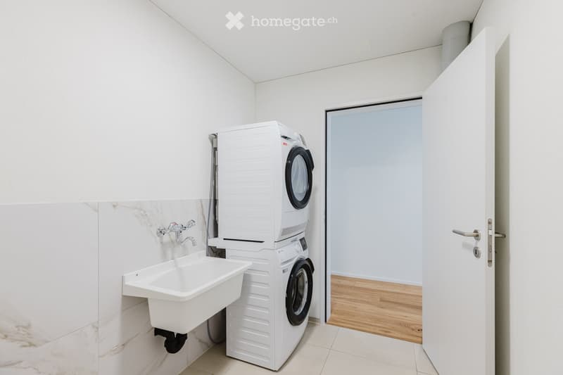 Modern bathroom with white tiled walls, a white sink, and a stacked washer/dryer unit. The floor is tiled, and an open door leads to another room with wooden flooring.