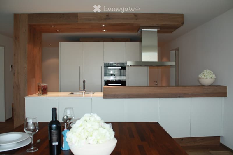 Modern kitchen with white cabinets, wooden countertops, stainless steel appliances, and a central island. Decor includes a vase of white flowers, wine bottle, and wine glasses on the table.