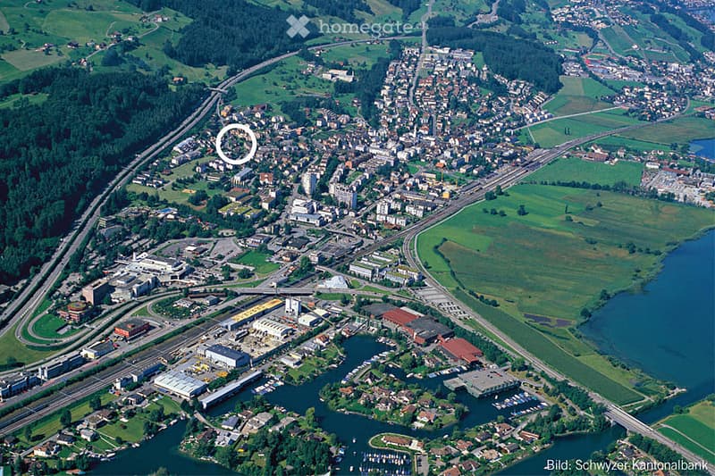 Aerial view of a town with a mix of residential and commercial buildings, surrounded by green fields and a body of water. Roads and highways are visible, along with a forested area on one side.