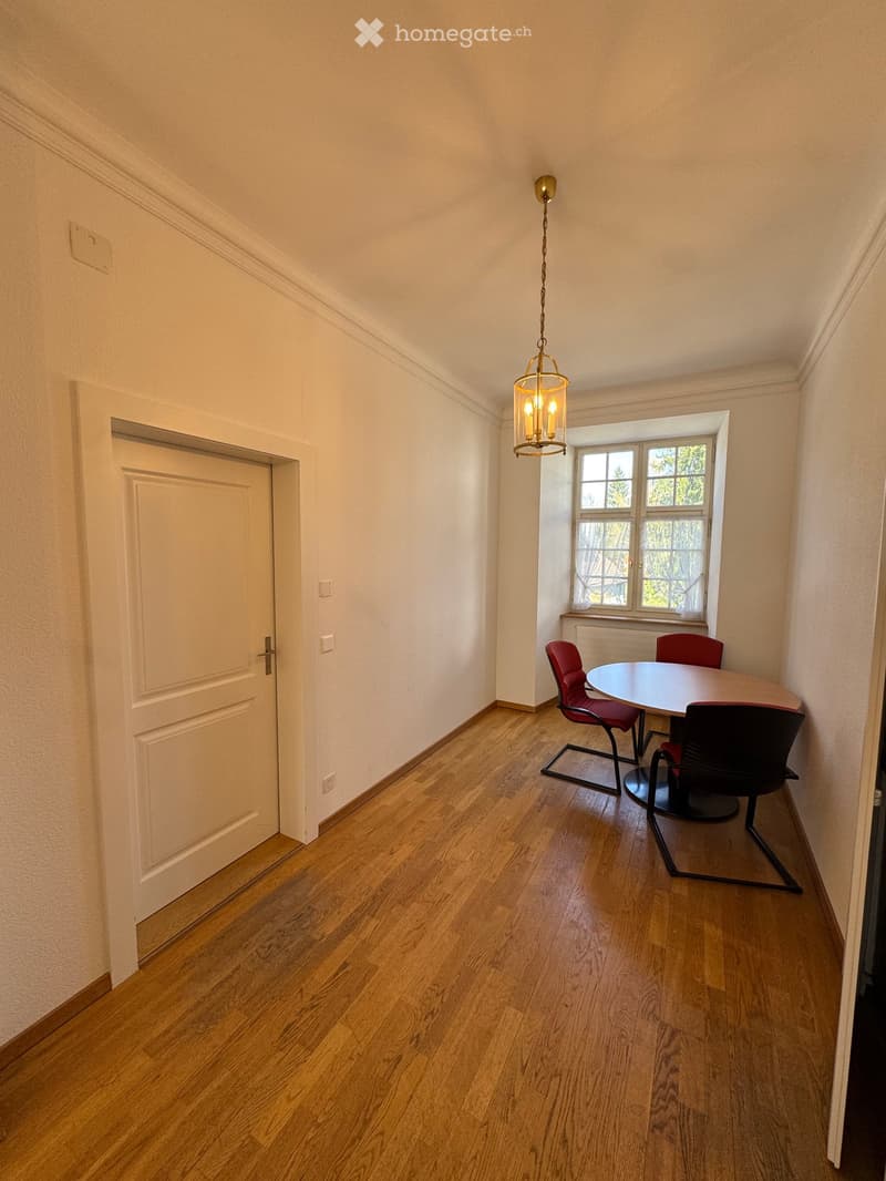 A small, unfurnished room with wooden flooring, a white door, and a window with a view of greenery. A round table with four chairs is placed near the window.