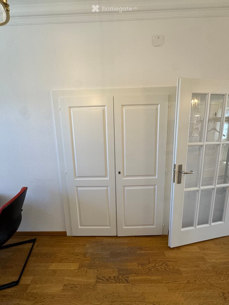 Empty hallway with wooden flooring, white double doors, and a red chair partially visible on the left side.