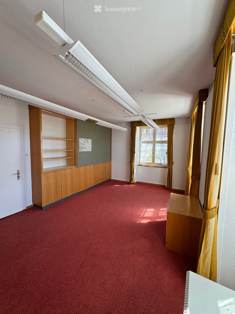 A furnished room with red carpeting, a wooden cabinet, and yellow curtains on the windows.