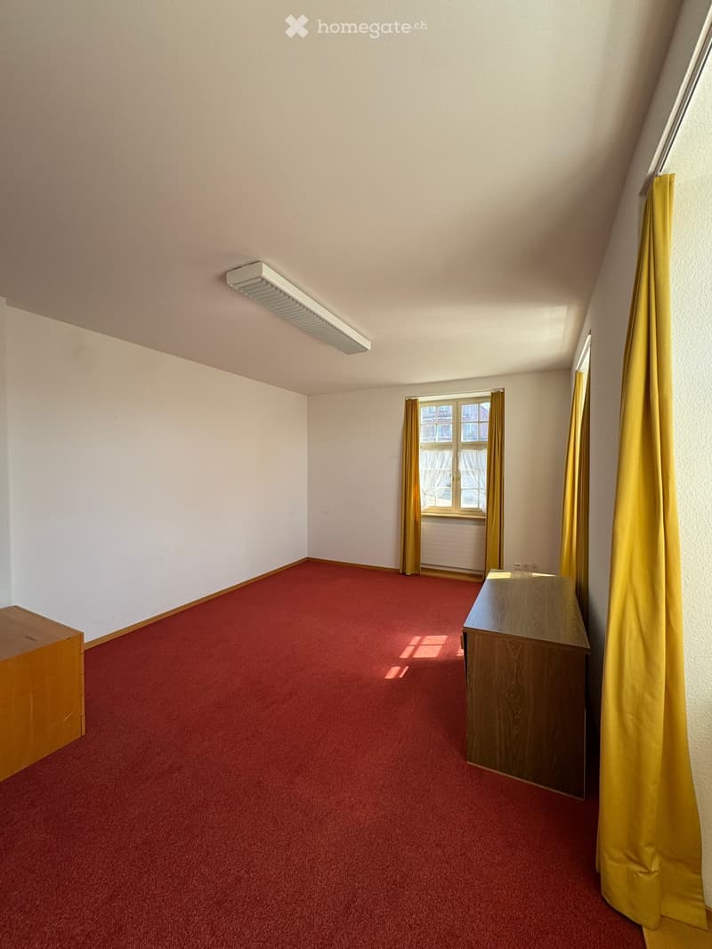 Empty room with red carpet, white walls, and a window with yellow curtains. A small wooden desk is positioned near the window.