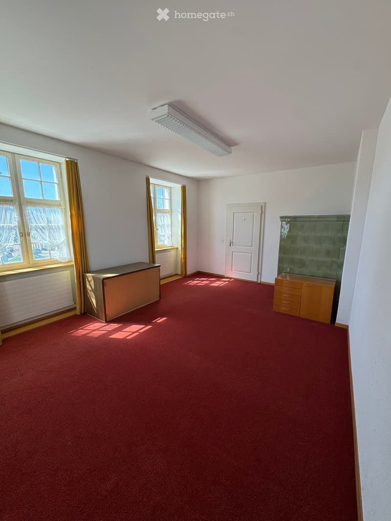 Empty room with red carpet, white walls, and large windows. Contains a small wooden cabinet and a table near the window.