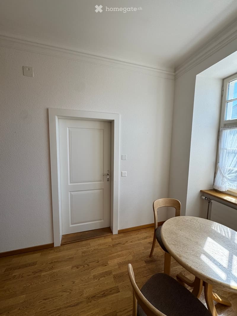 A small, unfurnished room with wooden flooring, a white door, a window with a curtain, and a small table with a chair.