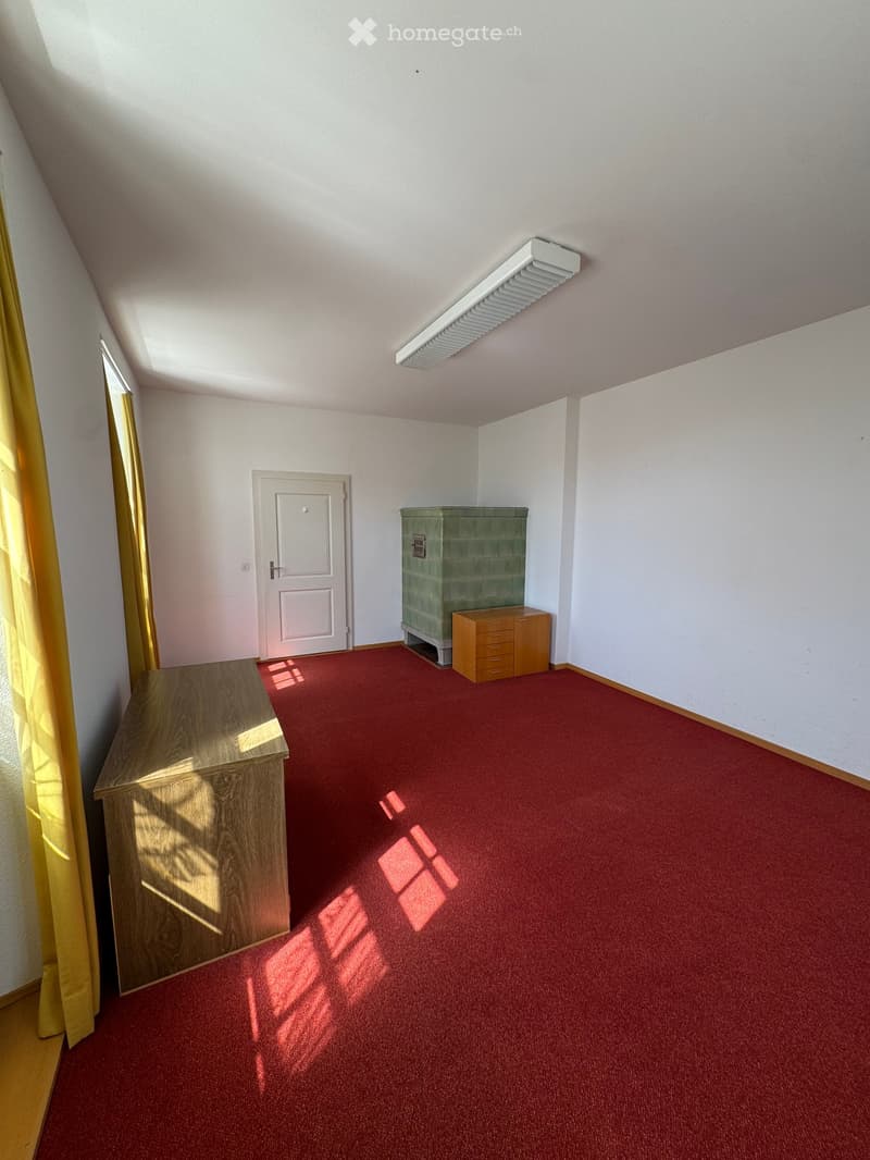 Empty room with red carpet, white walls, and wooden furniture including a cabinet and a drawer unit. A door is visible, and sunlight streams in from a window with yellow curtains.