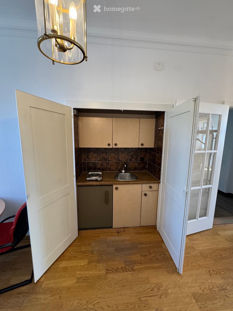 A small, well-lit kitchen with wooden flooring, cream-colored cabinets, a stainless steel sink, a small stove, and a dishwasher. A red chair is visible near the entrance.