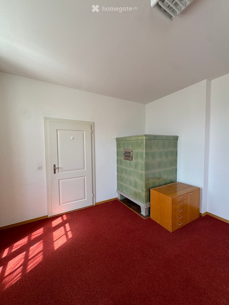 A small room with a red carpet, a white door, a green tiled fireplace, and a wooden cabinet with drawers.